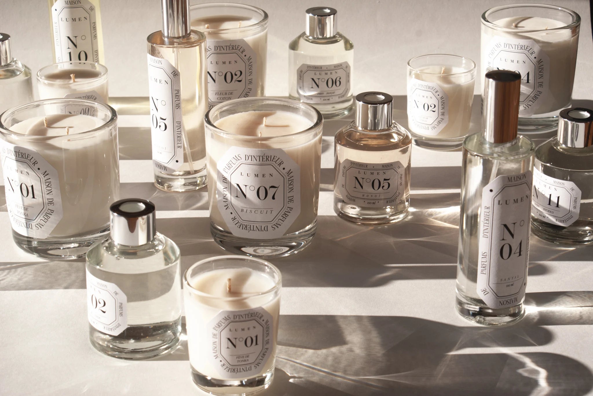 Photo qui présente une table blanche sur laquelle sont disposés de manière aléatoire des bougies parfumées LUMEN 180g, des bougies parfumées 70g, des vaporisateurs et des diffuseurs. L’éclairage met en évidence la qualité luxe des produits.