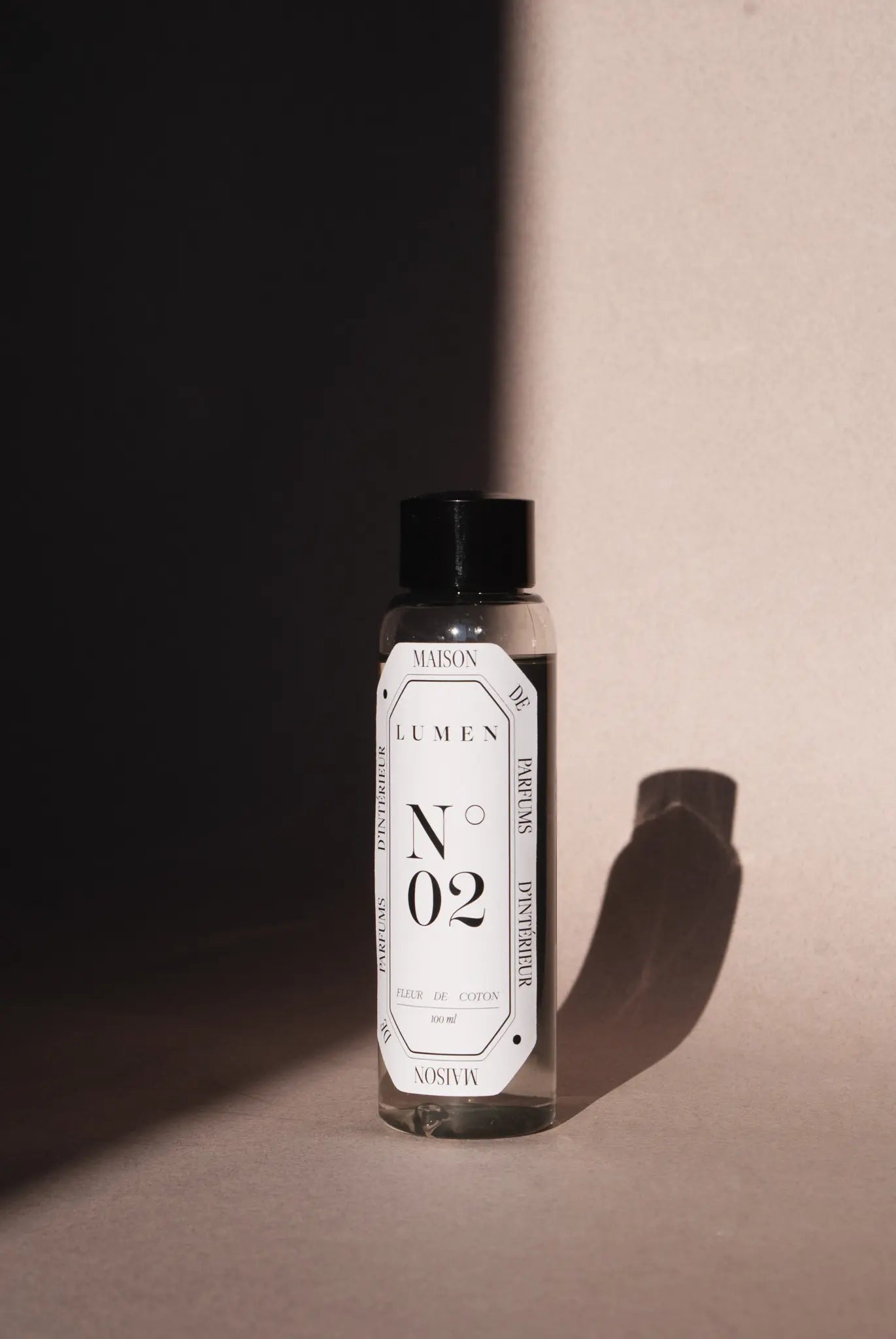Photo d'une recharge de parfum pour diffuseur et vaporisateur LUMEN. La photo est centrée sur la recharge qui est sur fond beige. Un jeu d'ombre met en avant le produit. L'étiquette indique LUMEN N°02 fleur de coton.