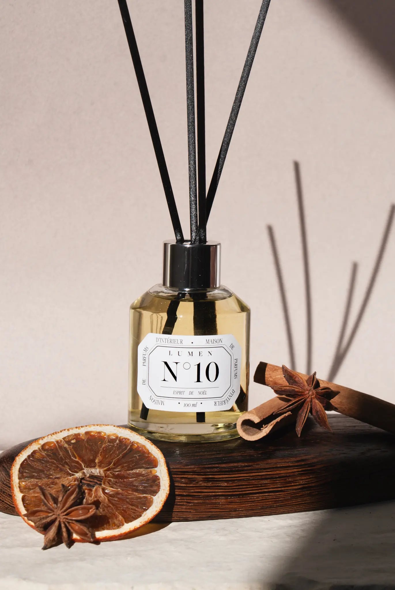 Photo d'un diffuseur à batons LUMEN N°10 esprit de noel. Au centre le diffuseur avec 4 batons en fibre noir très élégants, il est posé sur un plateau en bois, à gauche une tranche d'orange et une fleur d'anis, à droite deux batons de cannelle.