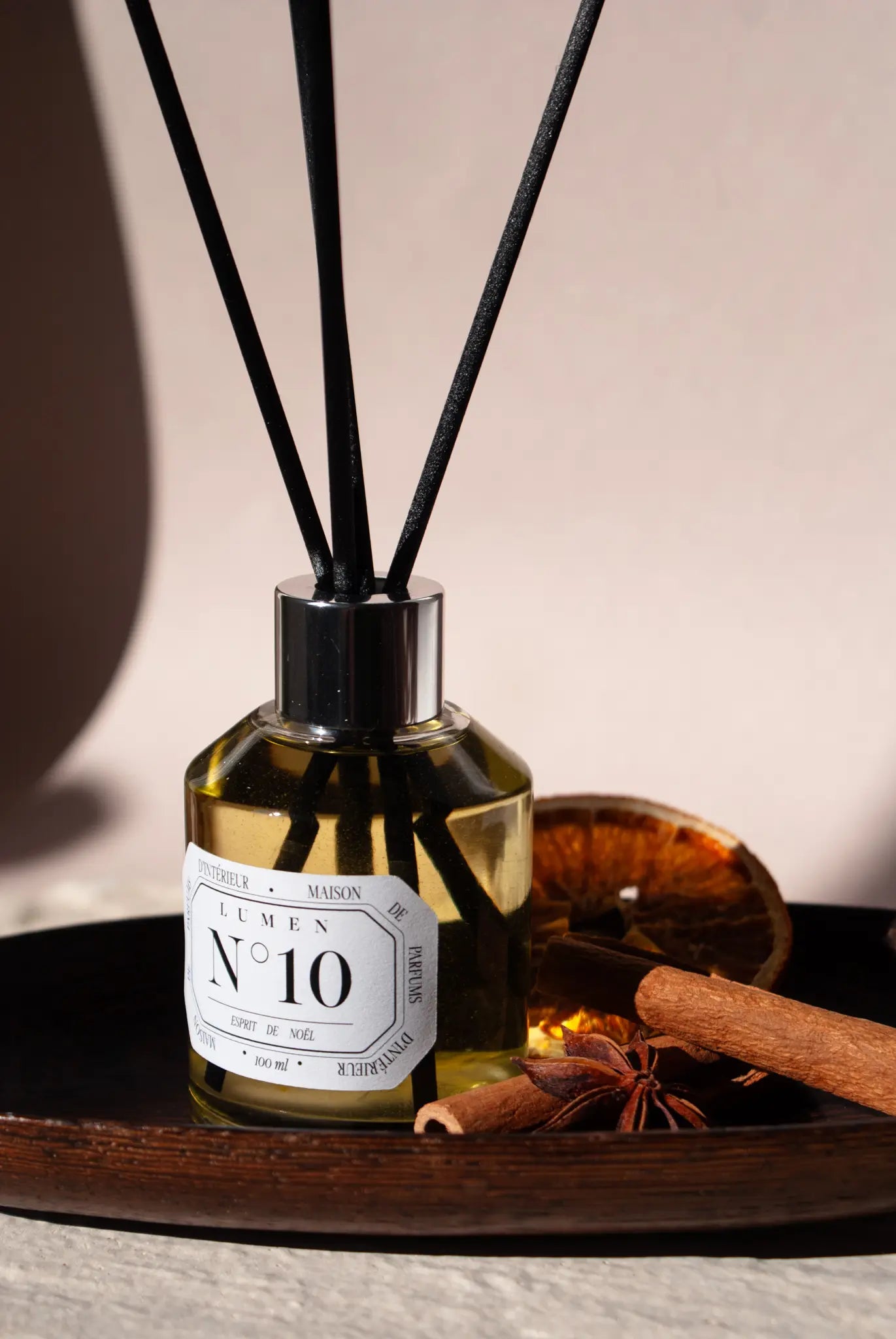 Photo d'un diffuseur lumen N°10 esprit de noel. Il est posé sur un plateau en bois, au dessus 4 batons en fibre noir. AA droite une tranche d'orange séchée, deux batons de cannelle et une fleur d'ani.