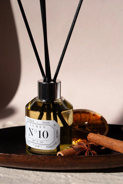 Photo d'un diffuseur lumen N°10 esprit de noel. Il est posé sur un plateau en bois, au dessus 4 batons en fibre noir. AA droite une tranche d'orange séchée, deux batons de cannelle et une fleur d'ani.